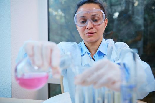 African American female chemist scientist, pharmacologist pouring pink liquid substance from glass laboratory flask into test tubes, while working on new drug in science pharmaceutical laboratory