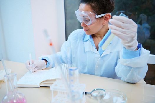 Multiethnic woman scientist, pharmacologist, researcher in a white lab coat and goggles writes in a notebook the results of experiments carried out in test tubes in a clinical research laboratory