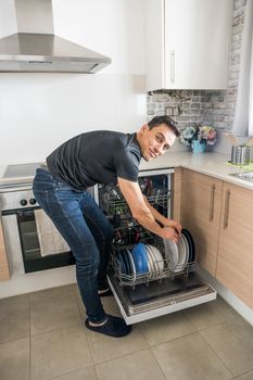 Man taking dishes out of the dishwasher in the kitchen and looking at camera smiling. Full body.