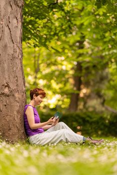 Side view of androgynous female in casual clothes sitting on lawn near tree and using tablet on sunny summer day in lush park