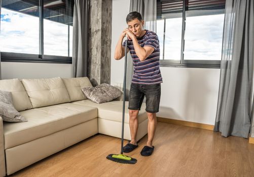 Sad and pensive man sweeping up his living room with casual clothes and slippers. Full body.
