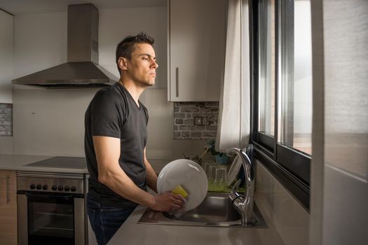 Thoughtful man washing dishes in the kitchen and looking through the window. Medium shot.