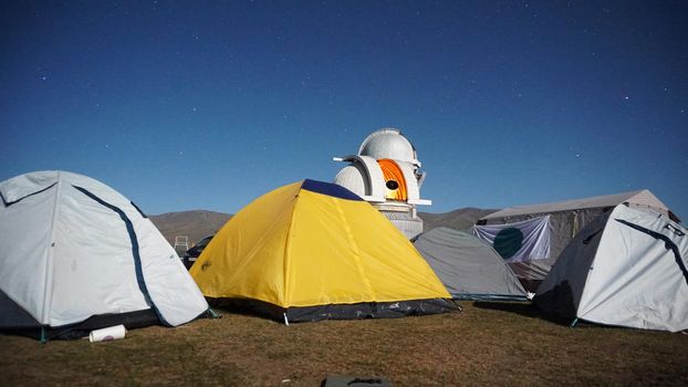 A camp with colored tents near the observatory. The Perseid starfall. A large teleskom watches the starry night sky. Meteorites and planes fly by, leaving traces in the sky. The stars are burning.