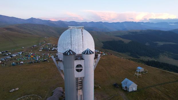 Two large telescope domes at sunset. Drone view of Assy-Turgen Observatory. Beautiful red sunset. Green hills and clouds. Tourists watch the sun. There is a large tent camp and cars nearby. Kazakhstan