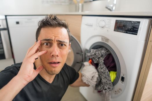 Man putting clothes in the washing machine, in the kitchen, worried because he has to wash a lot of clothes. Close up.