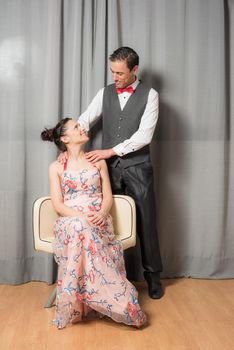 Portrait of smiling upper class marriage. The woman, seated in an elegant flowered dress. The man, standing, in an elegant suit. Full body.