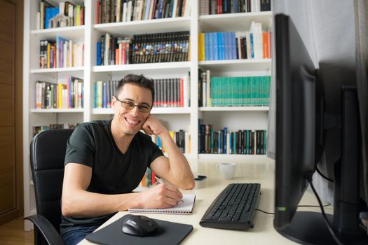 Smiling and happy man in his room sitting at the computer looking at camera, wearing a shirt. Mid shot.