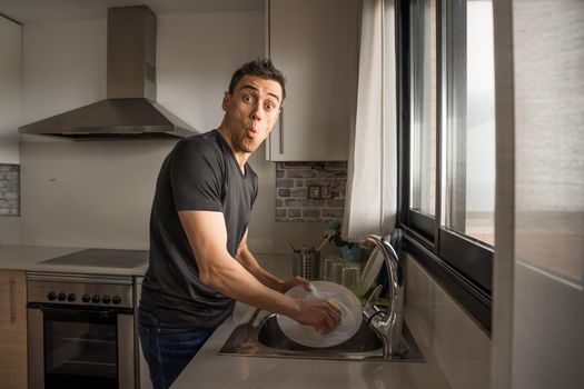 Man washing dishes in the kitchen next to a window making a funny face. Medium shot.