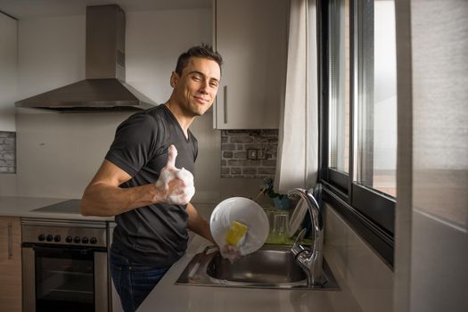 Smiling and happy man washing dishes in the kitchen next to a window. Medium shot.