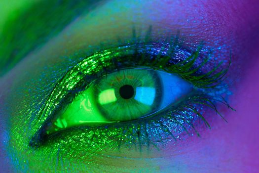 Extreme close up of female eye iris under neon light. Woman with beautiful makeup, glitter shadows and false lashes. Girls green eye. Nightlife, night club concept. High quality photo
