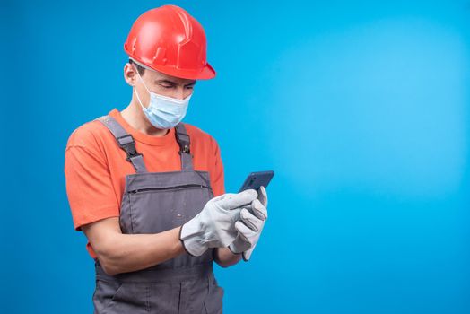 Man in builder uniform and gloves with face mask and hardhat watching video in social media on cellphone against blue background during pandemic