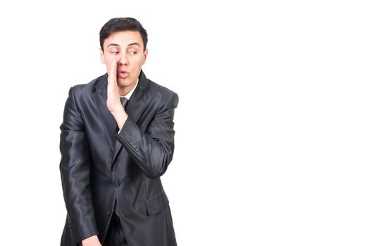 Positive man in elegant costume showing gesture with hand imitating whispering secret standing against white background
