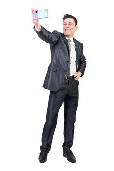 Full length of happy male entrepreneur in formal suit taking self portrait on smartphone while standing with hand on waist against white isolated background