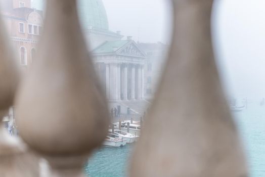 View through stone bridge on boats moored on rippled canal near aged historic building with columns in hazy day in Venice