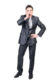 Full body of astonished young male manager in formal clothes touching cheek and looking at camera with opened mouth while standing against white background with hand on waist