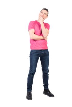 Full body of optimistic male in casual wear touching chin while looking away dreamily against white background