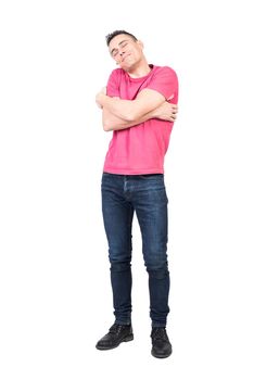 Full length of positive male in jeans and pink t shirt embracing shoulders and smiling with closed eyes while feeling love against while background
