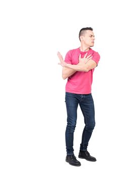 Full length of young serious guy with dark hair in casual clothes touching hearts and showing stop gesture with closed eyes isolated on white background