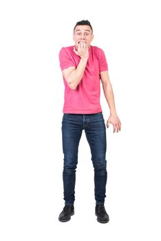 Full body of young nervous male with dark hair in t shirt and jeans biting nails and looking at camera against white background