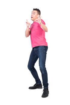 Full body of young funny male model with dark hair in pink t shirt and jeans looking and pointing at camera against white background