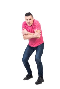 Full body of dissatisfied young male with dark hair in pink t shirt and jeans rubbing arms and looking at camera in cold studio
