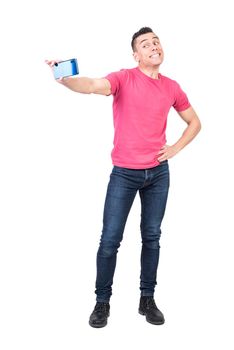 Full body of confident positive young male with dark hair in t shirt and jeans smiling happily while taking selfie on smartphone standing against white background with hand on waist