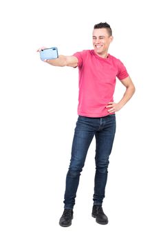 Full length of self assured young happy male with trendy haircut in casual outfit standing against white background with hand on waist and taking selfie on smartphone