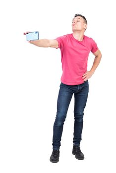 Full length of funny young guy with dark hair in t shirt and jeans pouting lips and taking selfie on mobile phone standing in studio with hand on waist