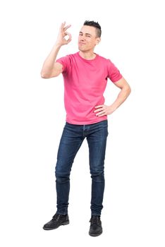 Full body of content young confident male model with stylish haircut in casual clothes showing OK gesture and looking at camera while standing against white background with hand on waist