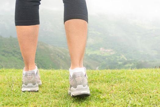 Back view of crop anonymous female tourist wearing sneakers standing on grassy hill and admiring picturesque view of green misty valley