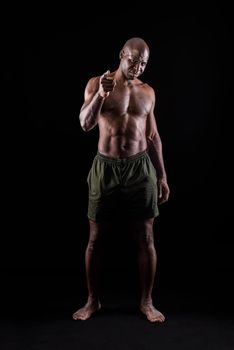 African american muscular man standing and pointing at camera with a confidence expression. Bodybuilder with bare torso and wearing shorts on a black background.