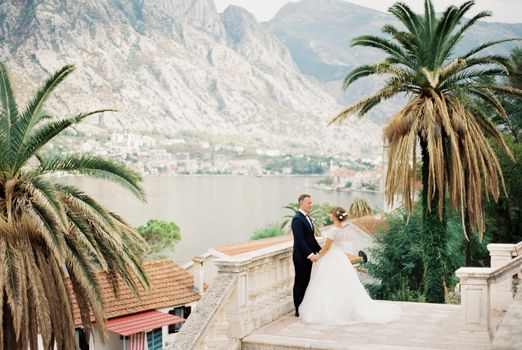 Bride and groom stand holding hands on the observation deck overlooking the sea and mountains. High quality photo