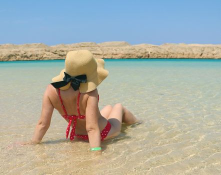 girl sunbathes in the water at the edge of the sea. In a swimsuit and hat. Blue sky and clear water, white and clean sand, mountains in front. A bright sunny day. Vacation concept by the sea, travel