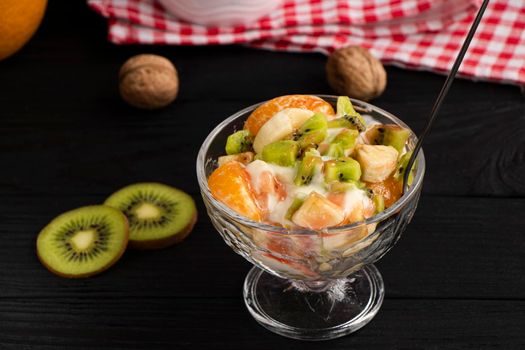 fruit salad in a glass bowl. Kiwi, banana and tangerine with yoghurt and fruit topping. A healthy, vitamin-rich snack. Healthy nutrition concept. Black wood background. High quality photo