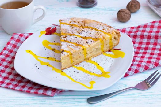 a piece of apple pie on a plate. Decorations with powdered sugar, cinnamon and topping. On a checkered kitchen napkin, with a fork and a cup of tea on a wooden table. High quality photo