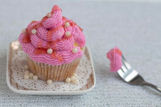 a cupcake beautifully decorated with pink cream and sugar decor stands on a square saucer on a light wooden background. A piece of muffin on a fork nearby. Sugar beads and fruit topping