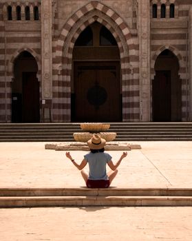 a girl in a straw hat sits with her back in a meditate pose before entering the temple. High quality photo. Yoga Concept. Hand Woman Practicing Lotus Pose