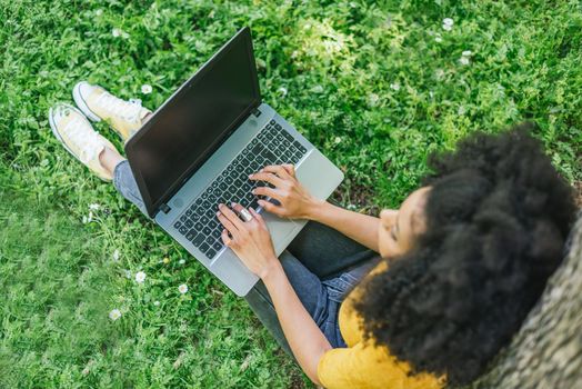 Afro woman typing on a laptop in a garden. Detail of hands on keyboard. Selective focus.