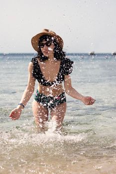 girl in a swimsuit and a straw hat in the sea. Splashing water. Emotion of joy. girl playing in the river. A girl raises her hands up in the water and splashes water drops