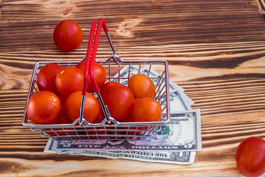 shopping basket filled with small cherry tomatoes. Stands on dollar bills. The concept of buying vegetables from farmers online. Burnt wood background. High quality photo