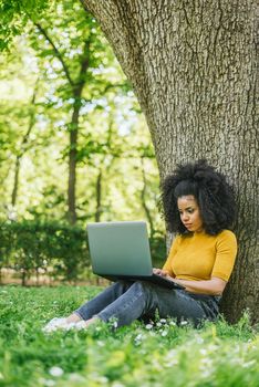 Beautiful afro woman typing on a laptop in a garden. Side view at grass level.