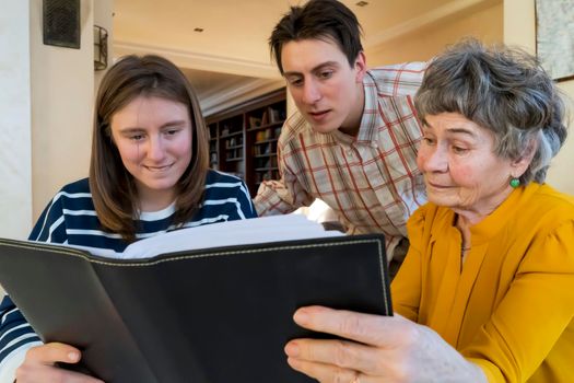 Family smiles happily and spends time together at home, young girl and a man look at photo album with their grandmother, help an elderly woman remember her youth, visit and take care of grandparents.