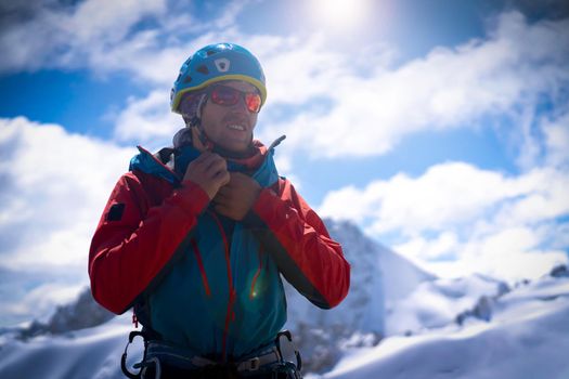 A man in special climbing equipment, in a membrane jacket, in a helmet and in sunglasses is preparing for ski touring, extreme sports, mountaineering in snowy mountains on a sunny day.