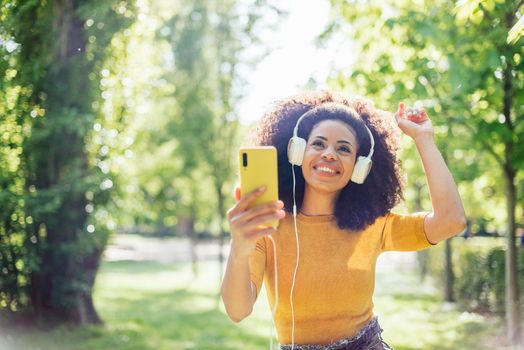 Pretty afro woman listens to music with headphones dancing in a garden. Selective focus.