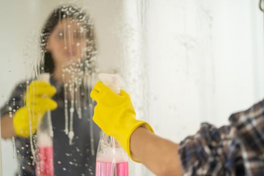 A young girl is cleaning the bathroom, applying detergent with a spray and washing the mirror with a sponge in yellow gloves on her hands. Woman taking care of the cleanliness of her home.