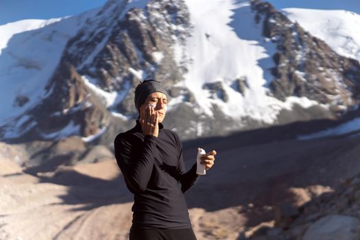 A young man applies sunscreen to his face in the highlands, in the background the peaks of the mountains are covered with snow. The traveler takes care of his skin while climbing.