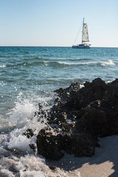 Seascape with waves breaking on the shore against some rocks, and a boat in the background.