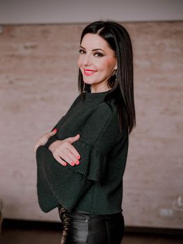 Portrait of adult beautiful mature woman standing on brown wall background. Lady in warm green sweater and leather pants with charming makeup and black hair smiling to camera.