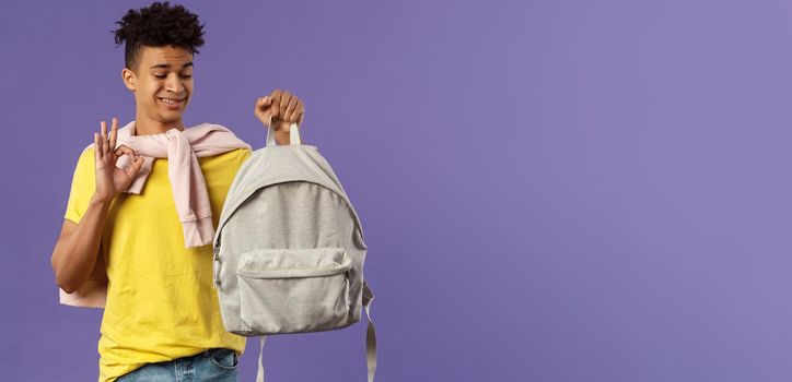 Portrait of young awkward guy, student receive gift new backpack from mom, being displeased with it grimacing but say okay, not bad, looking at rucksack standing purple background.