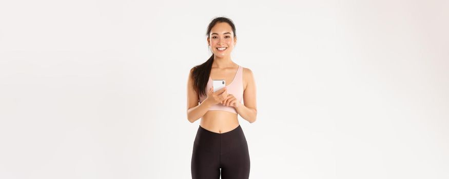 Sport, technology and active lifestyle concept. Smiling satisfied asian fitness girl, sportswoman holding smartphone, using running tracker app, checking heartrate during workout, white background.